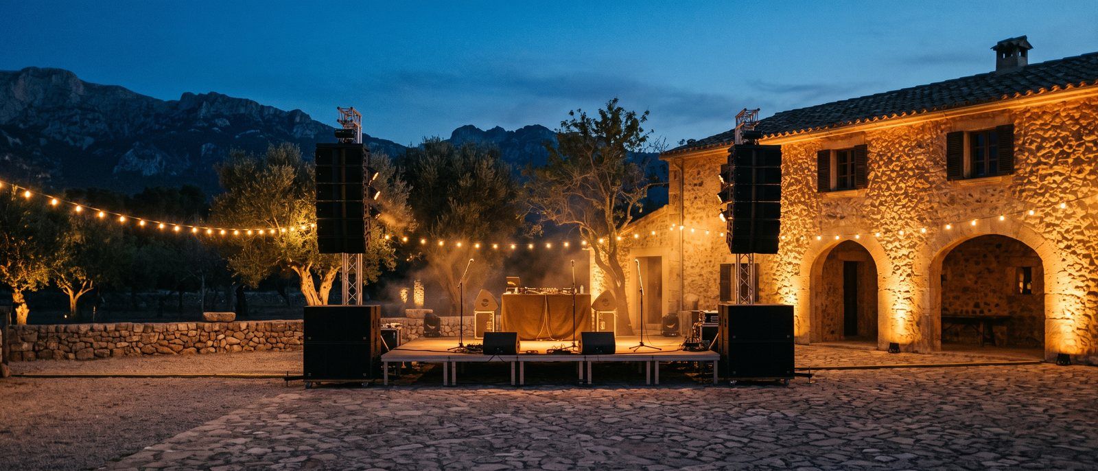 Gepflasterter Innenhof einer mallorquinischen Finca in der Dämmerung mit zwei Lautsprecher-Stacks neben einer Holzbühne