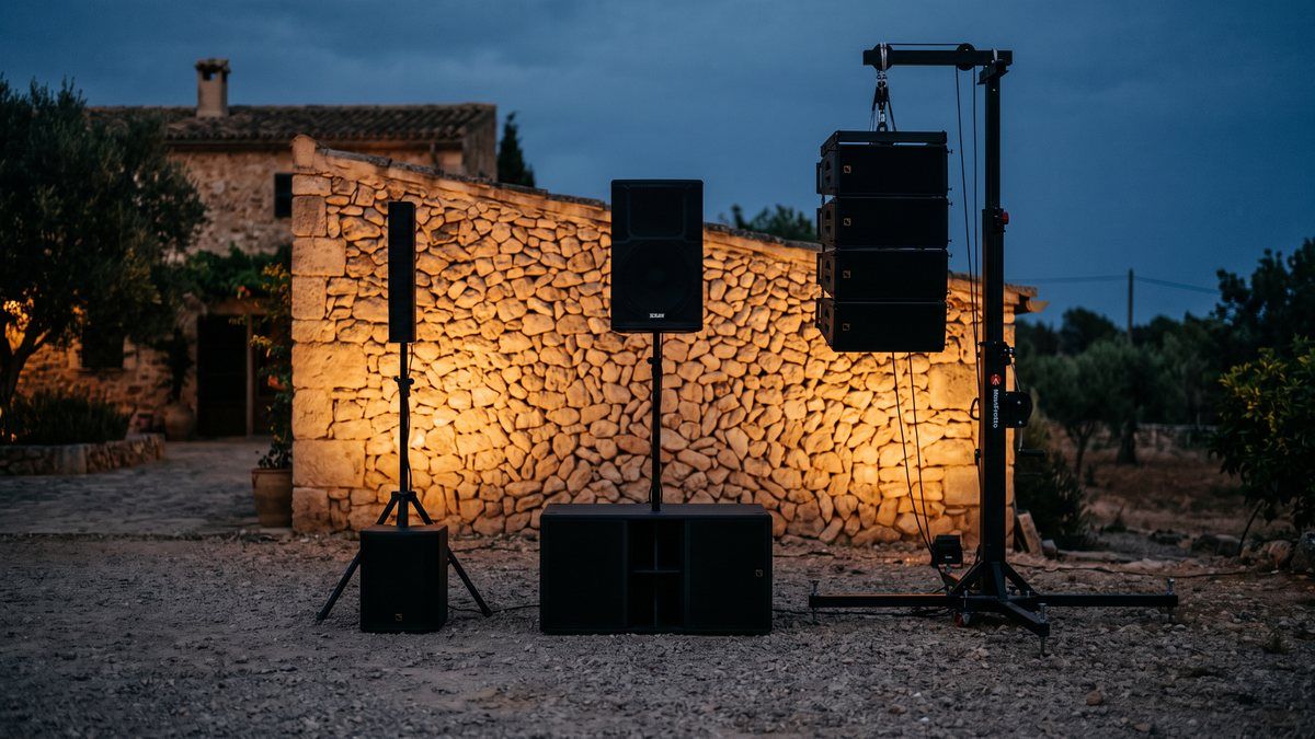 Three sizes of PA system lined up on gravel in front of a Mallorcan stone wall: portable column, stacked top over subwoofer, and a line array flown from a lifting tower