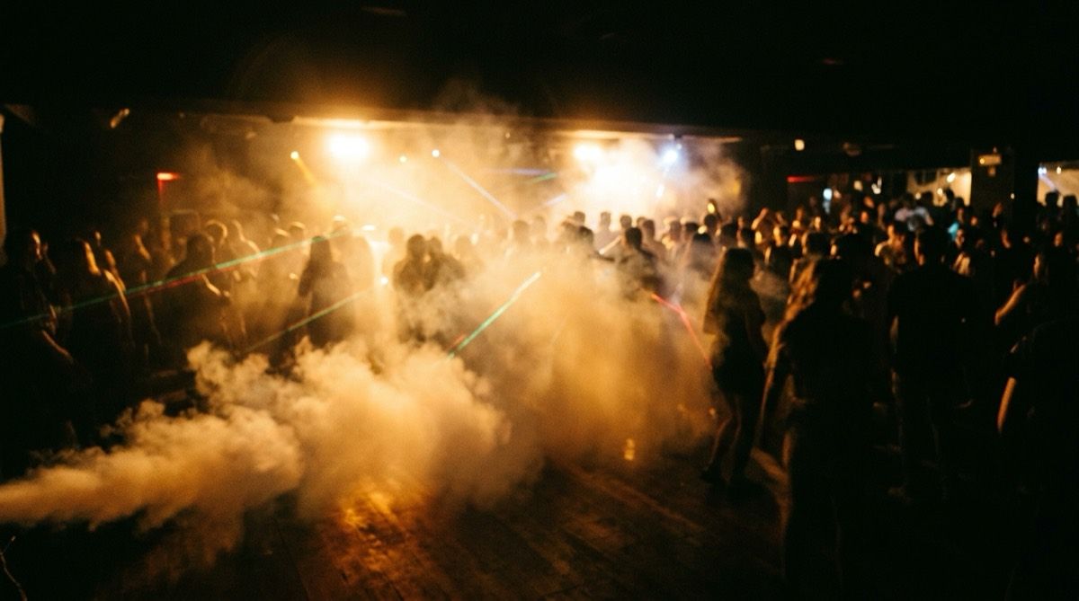 A dance floor covered in low haze, cut through by strobe bursts and warm-toned laser lines